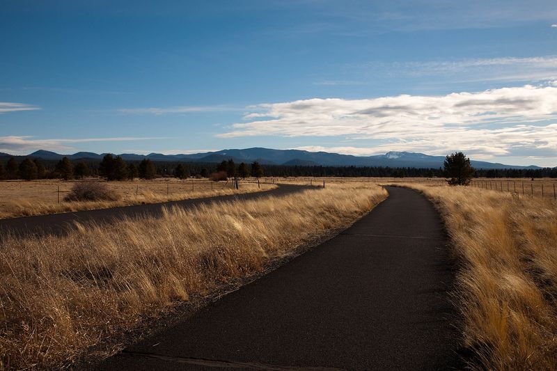 Paved Sunriver bike path running through golden meadow grass toward distant mountains
